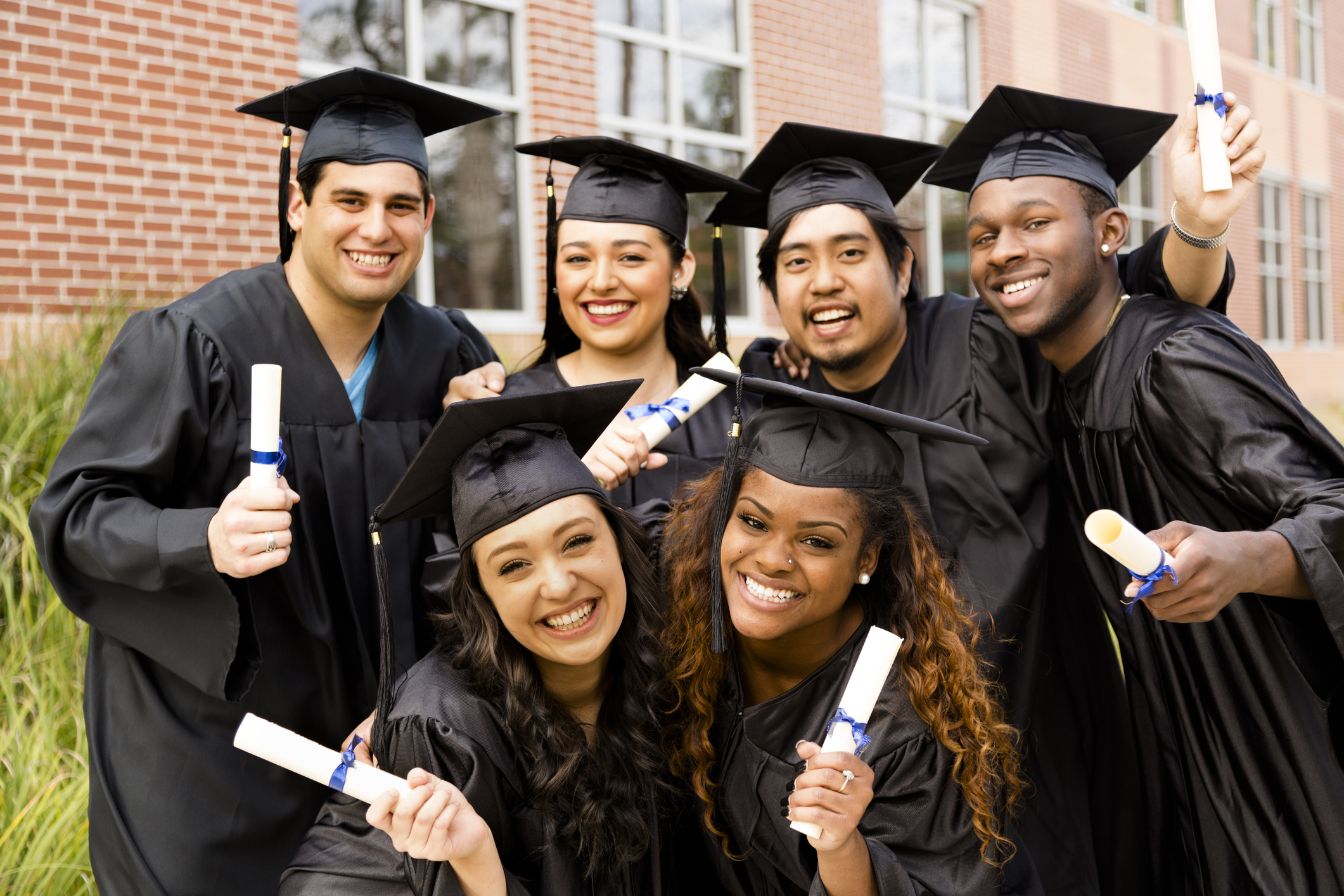 Diverse group of friends dressed in cap and gowns excitedly show off diplomas after college graduation. School building background.