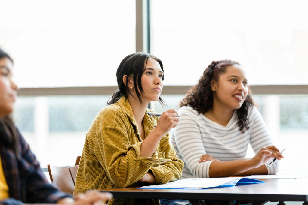 A diverse group of young adult female college students have serious looks on their faces as they watch a video in their class.